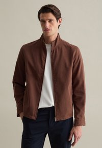 Braune Zip-Jacke mit Stehkragen, aus glattem Baumwollstoff gefertigt. Kombiniert mit einem weißen T-Shirt und dunkelblauen Hosen. Einfaches Design.