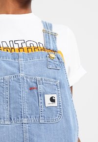 Pantalones de mezclilla de tono azul claro con tirantes ajustables, bolsillo frontal y parche con el logo de Carhartt. Complementados con una camiseta blanca con texto impreso.