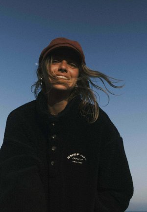 Kobieta w bordowej czapce i czarnej kurtce z logo "Surf Inn" uśmiecha się, a jej włosy powiewają na tle jasnego, zmierzchowego nieba.