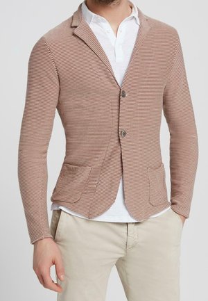 Mężczyzna ubrany w beżowo-biały marynarkę w paski na dwa guziki, białą koszulkę polo oraz jasnobeżowe spodnie, stojący na jednolitym tle.