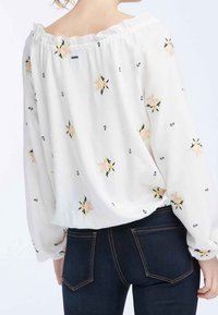 Blusa bianca senza spalline con ricami floreali, colletto elastico e maniche lunghe a palloncino, abbinata a jeans in denim scuro.