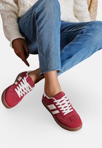 Baskets en suède rouge avec des accents et des lacets blancs, dotées d'une semelle en caoutchouc marron. Associées à un jeans bleu effiloché et un pull en tricot blanc.