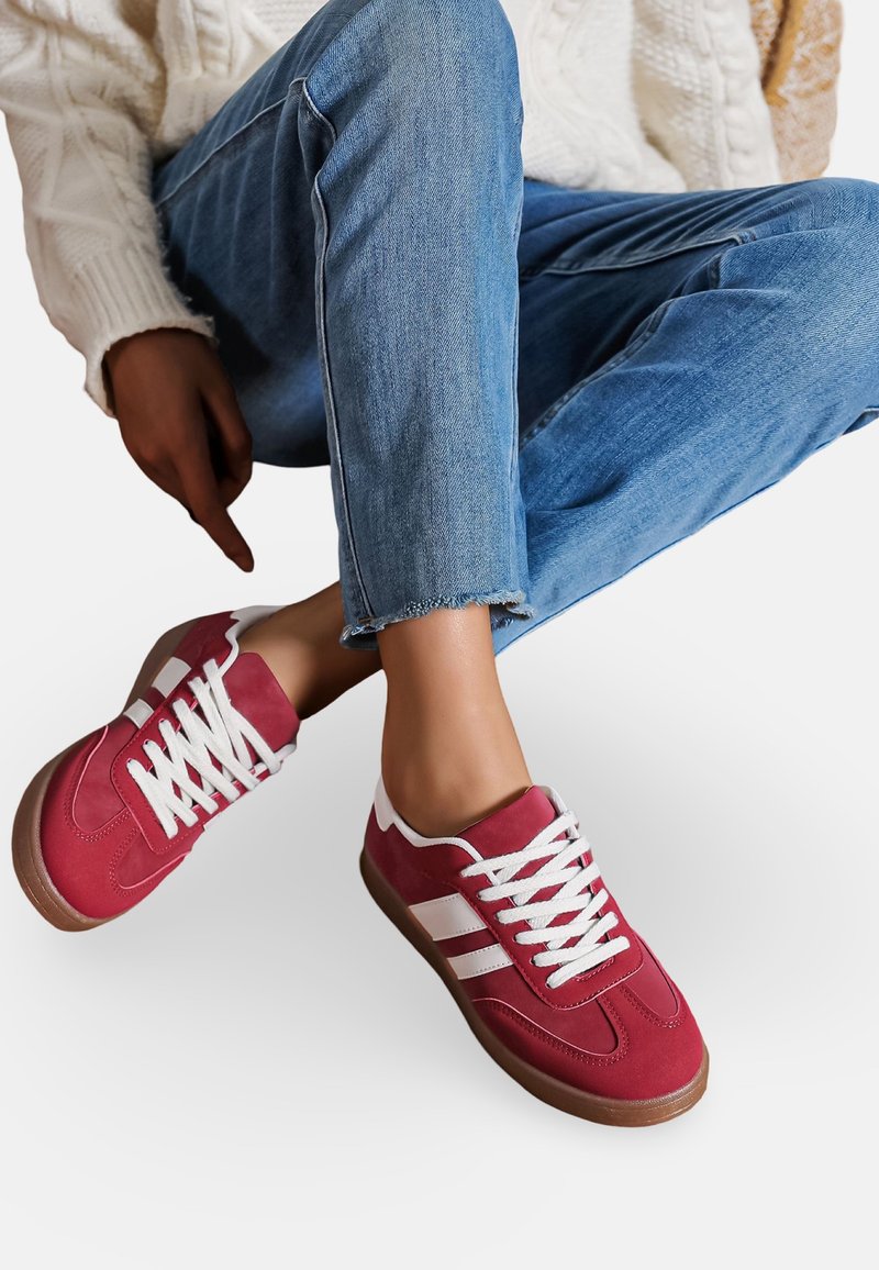 Baskets en suède rouge avec des accents et des lacets blancs, dotées d'une semelle en caoutchouc marron. Associées à un jeans bleu effiloché et un pull en tricot blanc.