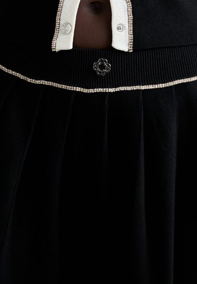 Svart skjørt med plisserte detaljer, elastisk liv, og en kremfarget knapp-up topp med perledetaljer og blomsterdekorasjon i livet.