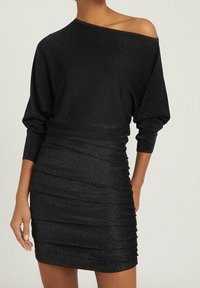 Kobieta w czarnej, błyszczącej, marszczonej mini sukience z szerokim dekoltem, odsłaniającym jedno ramię, stojąca z jedną ręką na biodrze.