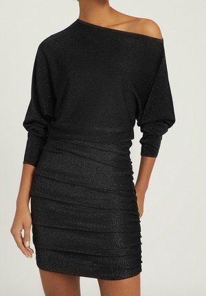 Femme portant une mini robe noire scintillante à fronces avec un grand col bateau découvrant une épaule, se tenant avec une main sur la hanche.