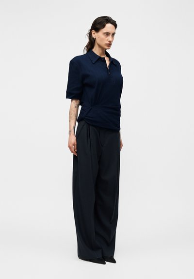 Γυναίκα με σκούρα μαλλιά φοράει μπλε σκούρο κοντομάνικο τοπ με γιακά και φαρδιά μαύρη παντελόνα, στέκεται μπροστά σε ανοιχτό μονόχρωμο φόντο.