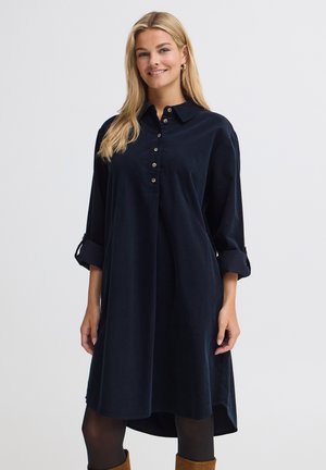 Femme blonde souriante, portant une robe bleu marine à manches longues boutonnée avec les manches retroussées, des collants noirs et des bottines marron.