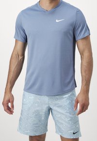 Nike urheilu paita vaaleansinisessä värissä, lyhyet hihat, V-pääntie. Yhdistetty vaaleansiniset kuviolliset shortsit, joissa on musta logo.