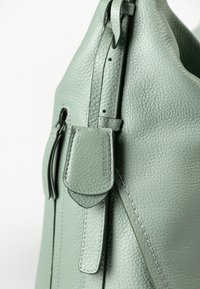 Primo piano di una borsa a tracolla in pelle verde salvia dalla texture evidente, con etichetta del marchio in rilievo e cuciture dettagliate su tracolla e pattina.