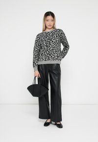 Pull gris à imprimé léopard noir, associé à un pantalon large en cuir noir et à un sac à main noir. Le modèle se tient devant un fond blanc.
