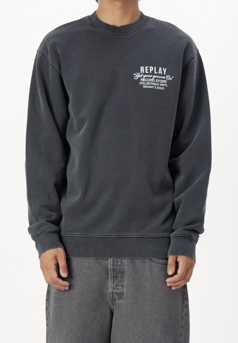 Personne portant un sweat-shirt gris foncé avec un texte blanc indiquant « REPLAY Get your groove On Record Store Collectable Vinyl Bought & Sold » sur la poitrine.