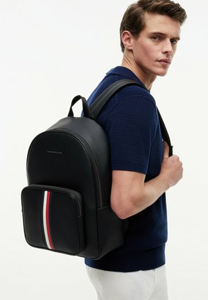Man wearing navy knit shirt and white pants carries black backpack with red, white, and blue stripe on front pocket against white background.