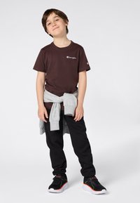 T-shirt di cotone marrone con logo, felpa grigia legata in vita, pantaloni felpati neri e scarpe da ginnastica nere e rosse. Sfondo neutro.