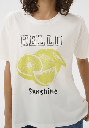 T-shirt blanc avec graphisme de tranches de citron jaune et texte "HELLO Sunshine", porté par une personne aux cheveux bruns bouclés.