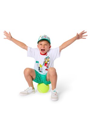 Jonge jongen die een pet en een cartoon T-shirt draagt, zittend op een grote groene tennisbal met opgestoken armen en open mond, uitdrukking van opwinding.