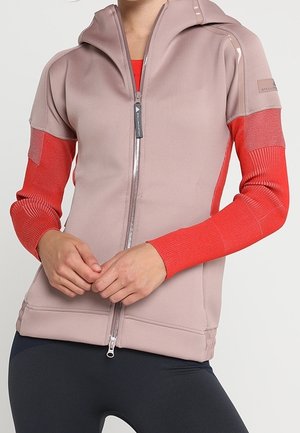 Žena v mikině na zip s béžovým tělem a červenými texturovanými rukávy, stojící s rukama sepnutýma v pase.