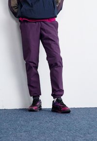 Purple lightweight athletic pants with an elastic waistband, paired with black and pink patterned sneakers, standing against a white wall.