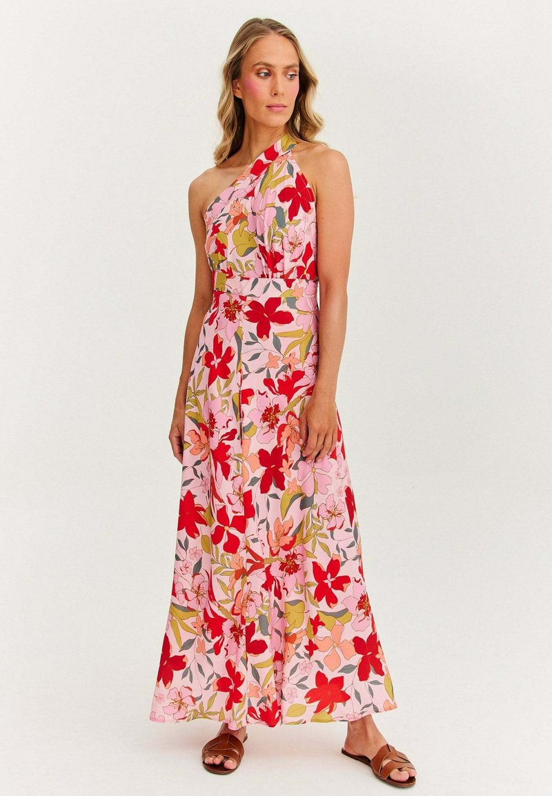 Žena oblečená v bezrukávových, jednoramenných ružových maxi šatách s červeným a oranžovým kvetinovým vzorom, doplnená hnedými plochými sandálmi.