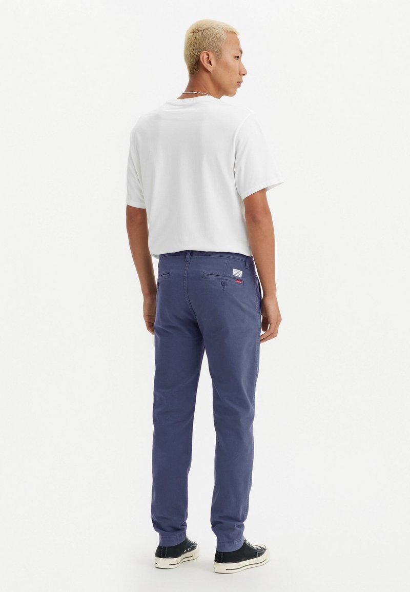 Chinos azul oscuro con bolsillos traseros y etiqueta. Combinados con una camiseta blanca y zapatillas negras de corte bajo.