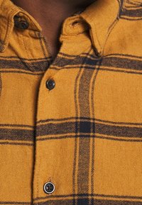 Chemise en flanelle jaune avec un motif à carreaux noir et gris. Elle possède un col à rabat et des boutons noirs sur la patte de boutonnage. Texture douce.