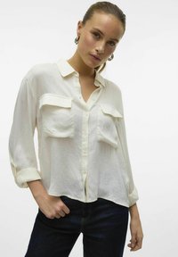 Camicia bianca leggera con bottoni, due tasche frontali, colletto classico e maniche arrotolate, abbinata a jeans in denim scuro.