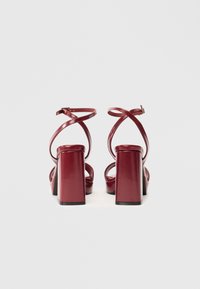 Sandalieri cu toc înalt de culoare burgundy, cu toc gros și bretele încrucișate la glezne; fabricate dintr-un material sintetic lucios, cu o textură netedă.