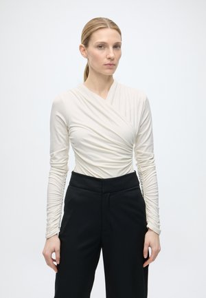 Donna con capelli biondi raccolti in una coda bassa, che indossa una maglietta bianca a maniche lunghe arricciata e pantaloni neri a vita alta, in piedi su uno sfondo semplice.