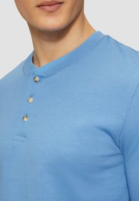 Šviesiai mėlynas ilgomis rankovėmis henley marškinėliai, pagaminti iš medvilnės, su grioveliu apykakle, trimis perlų mygtukais ant krūtinės ir prigludusia forma.