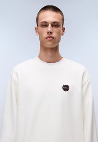 White sweatshirt featuring a round black and orange logo patch on the chest, made from a soft, smooth fabric with a relaxed fit.