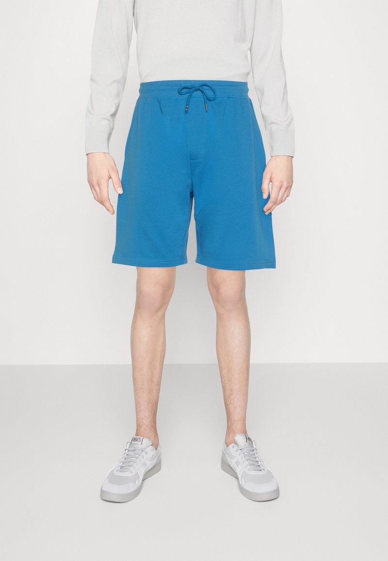 Personne portant un short bleu à cordon, une chemise blanche à manches longues et des baskets blanches, debout devant un fond clair uni.