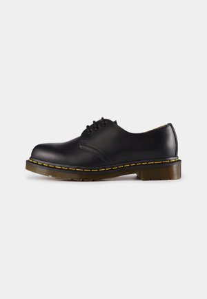 Zapato oxford de cuero negro con punta redonda, seis ojetes, costuras amarillas y una suela de goma gruesa y texturizada.