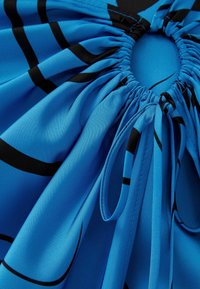 Sininen kangas, jossa on sileä pinta ja mustia abstrakteja viivoja. Kerroksellinen muotoilu ja solmittavat nauhat kaula-aukossa säätömahdollisuudelle ja tyylille.