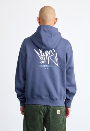 Homme au crâne rasé portant un sweat à capuche bleu Nike avec un texte et un logo graffiti blancs dans le dos, associé à un pantalon en velours côtelé vert.