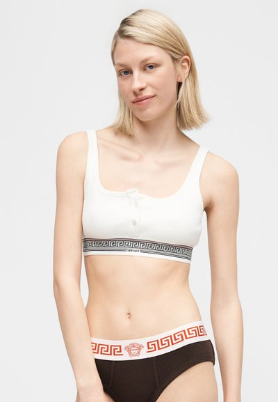 Γυναίκα με κοντά ξανθά μαλλιά που φοράει λευκό αμάνικο crop top και σκούρα εσώρουχα με ζώνη με ελληνικό κλειδί και λογότυπο Μέδουσας.