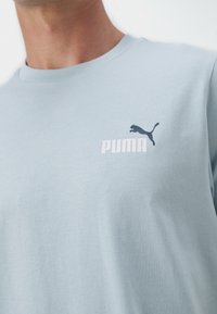 Camiseta de algodón azul claro con mangas cortas, que presenta un logo negro de Puma y texto blanco. Textura suave y cuello clásico tipo crew.