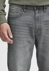 Jeans din denim gri cu un finisaj uzat, buzunare standard și cusături vizibile. Materialul are o textură netedă cu ușoare cute.