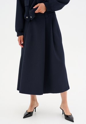 Navy midi skirt with pleats, made from a textured fabric. Paired with open-toe black heeled mules. Clean, minimal design.