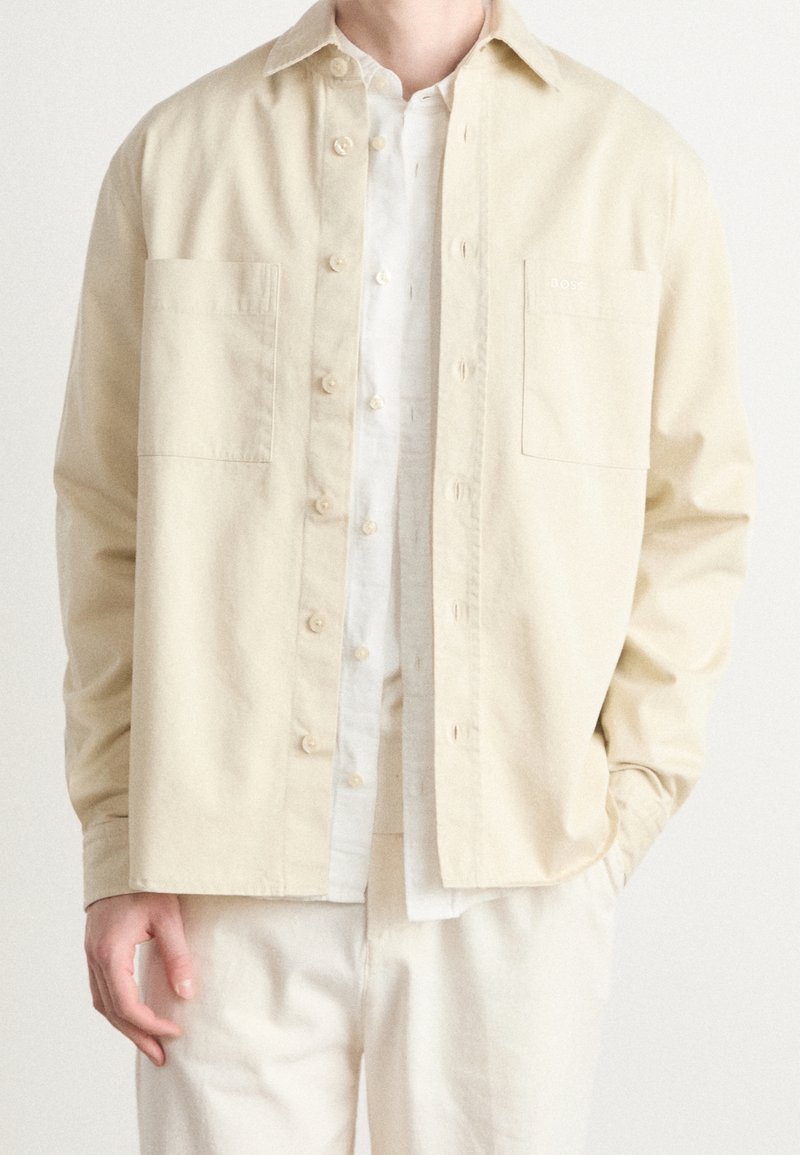 Homme portant une chemise beige clair boutonnée par-dessus une chemise blanche boutonnée avec un pantalon beige clair assorti, une main dans la poche.