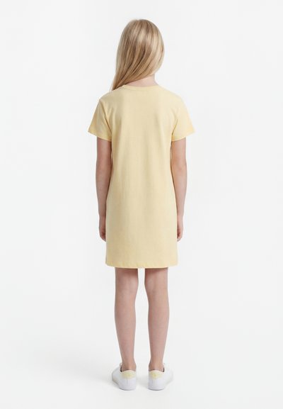 Παιδί με μακριά ξανθά μαλλιά, φορώντας κοντομάνικο κίτρινο φόρεμα και λευκά αθλητικά, στέκεται γυρισμένο με την πλάτη σε ένα απλό λευκό φόντο.