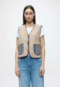 Μπεζ γιλέκο από φλις με πολύχρωμα κεντήματα στις τσέπες, μπλε περιθώριο και κούμπωμα με τογκλ. Φορεμένο πάνω από ένα λευκό πουκάμισο και τζιν.
