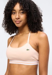 Νεαρή γυναίκα με σγουρά μαλλιά που φοράει ανοιχτό ροζ αθλητικό σουτιέν με λεπτές τιράντες και ένα μικρό πράσινο λογότυπο κροκόδειλου στο στήθος.