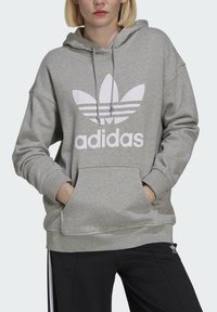 Pilkas gobtuvų džemperis, pagamintas iš minkštos medžiagos, su dideliu baltu Adidas logotipu ir dviem priekinėmis kišenėmis. Laisvas siluetas ir raukiniai ant rankovių.