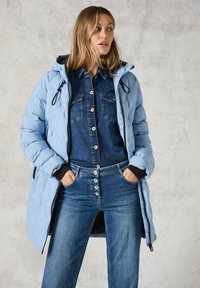 Ljusblå puffjacka med huva, svarta detaljer, matchad med en denimskjorta och högmidjade blå jeans. Texturerad, modern design.