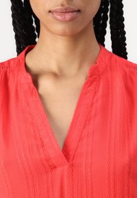 Blusa corallo con scollatura a V e dettagli di trama intrecciata, caratterizzata da sottili strisce e un tessuto morbido e leggero. Vista ravvicinata del collo.
