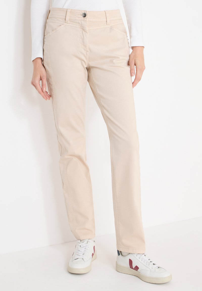 Pantalon beige à jambes droites porté avec des baskets blanches avec des accents bordeaux, associé à un haut blanc à manches longues, personne debout.