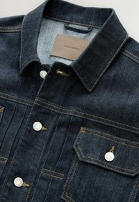 Tummansinisestä denimistä valmistettu takki, jossa on terävä kaulus, hopeiset napit, kaksi rintataskua ja nahkaleima, jossa lukee "DENIM SUPPLY."