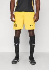 Gule fotballshorts med svarte detaljer, med en rund BVB-logo, kombinert med svarte knelengde sokker og svarte Puma-sko med teksturert design.