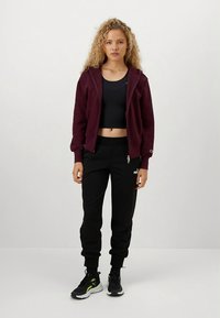 Camisola de capuz com fecho de correr em cor bordô sobre um top preto curto, combinada com calças de fato de treino pretas com logótipo, e ténis pretos com detalhes em neon.