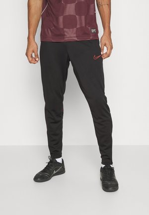 Schwarze Sporthose mit schmaler Passform, ausgestattet mit einem roten Nike-Logo auf dem linken Oberschenkel und kontrastierenden Nähten, kombiniert mit schwarzen Sportschuhen.